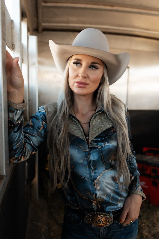 Marbled blue barrel racing shirt with gold lightning accents, glittery gold collar and cuffs, and a button-down front, worn by a woman with a cowboy hat.