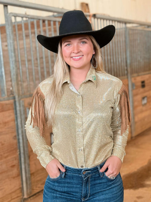 A young woman with long blonde hair wears a black cowboy hat and a shimmering gold fringe blouse while posing in a barn. She smiles confidently, holding onto a metal gate with both hands. She is dressed in dark blue denim jeans, and the rustic wooden walls of the barn serve as the backdrop. The outfit exudes Western style with a touch of glamour.